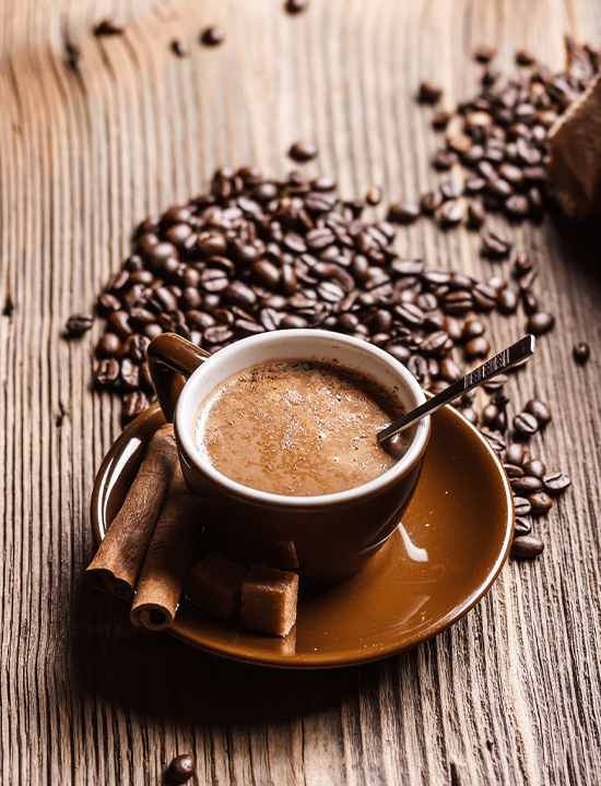 Rich, creamy coffee in a brown cup with a spoon, surrounded by coffee beans, cinnamon sticks, and sugar cubes on a wooden surface.